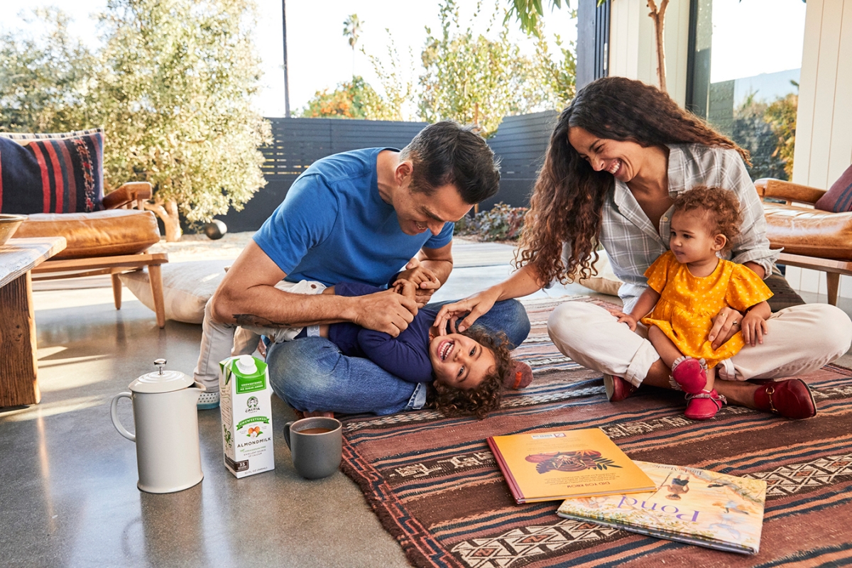 Family of four sitting on a patterned rug outside their house with story books, almond milk, and a french press coffee maker.