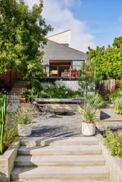 Modern house exterior with a multi-level terraced garden, concrete steps, and an elevated wooden deck under a bright blue sky.