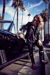 Blonde man in leather jacket leaning against a black classic car on a palm tree lined street by Justin Coit