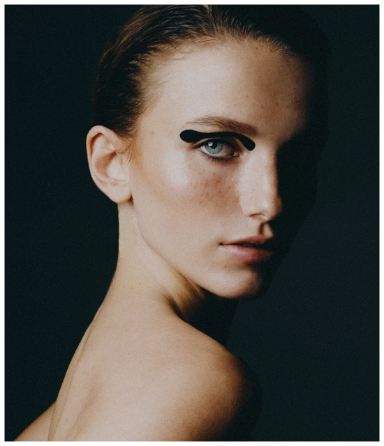 Portrait of a fair-skinned woman with graphic black eyeliner on a dark background, face angled to the side, with a bare shoulder visible.
