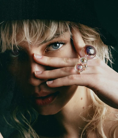 Close-up portrait of a blonde woman wearing a black hat, looking through her fingers while displaying an iridescent glass bubble ring.