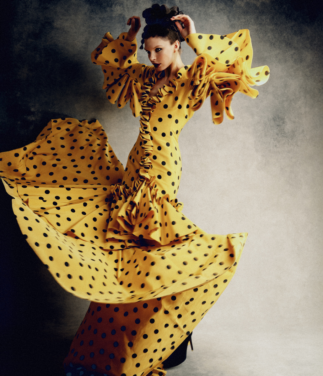 Elegant woman in motion wearing a yellow polka-dot dress with ruffled sleeves and a full skirt against a gray backdrop, fashion shoot.