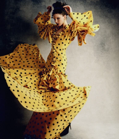 Elegant woman in motion wearing a yellow polka-dot dress with ruffled sleeves and a full skirt against a gray backdrop, fashion shoot.