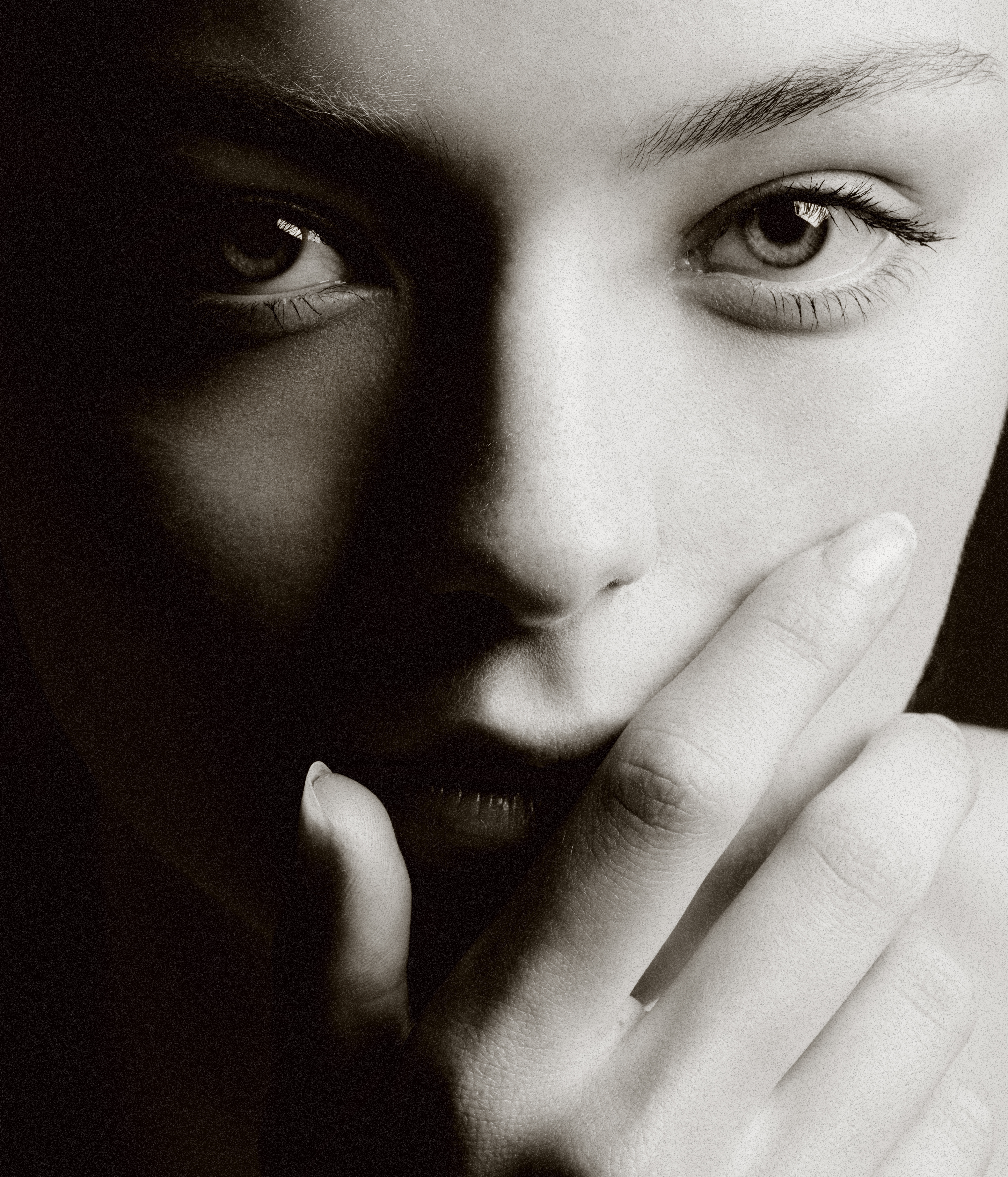 Close-up portrait of a young adult woman with hand near mouth, neutral expression, and studio lighting, in black and white.
