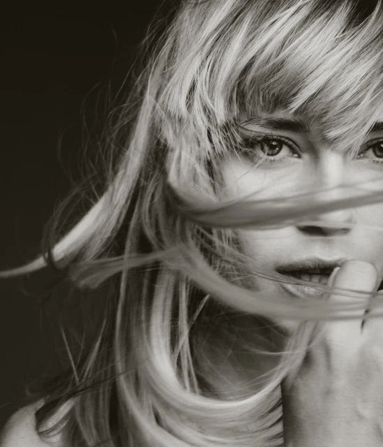 Close-up black and white portrait of a blonde woman with flowing hair covering part of her face, finger near lips.