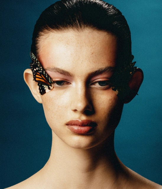 Close-up portrait of a young woman with butterfly decorations near her eyes against a blue background, showcasing makeup and beauty.