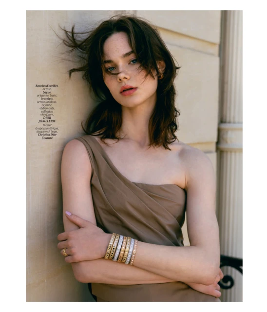 Portrait of a brunette woman in a tan dress with multiple bracelets, leaning against a textured wall in natural light.