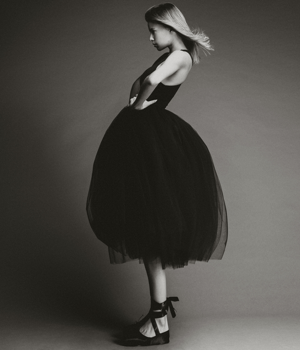 Young woman in a black dress and ballet shoes poses in a photo studio. The black and white image features a minimalist aesthetic and classic style.