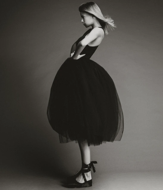 Young woman in a black dress and ballet shoes poses in a photo studio. The black and white image features a minimalist aesthetic and classic style.