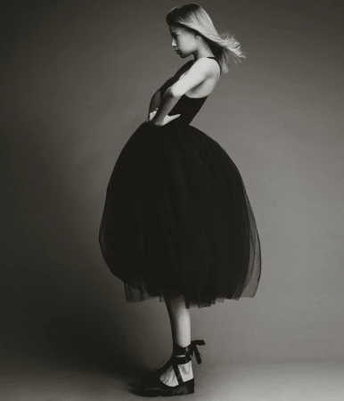 Young woman in a black dress and ballet shoes poses in a photo studio. The black and white image features a minimalist aesthetic and classic style.