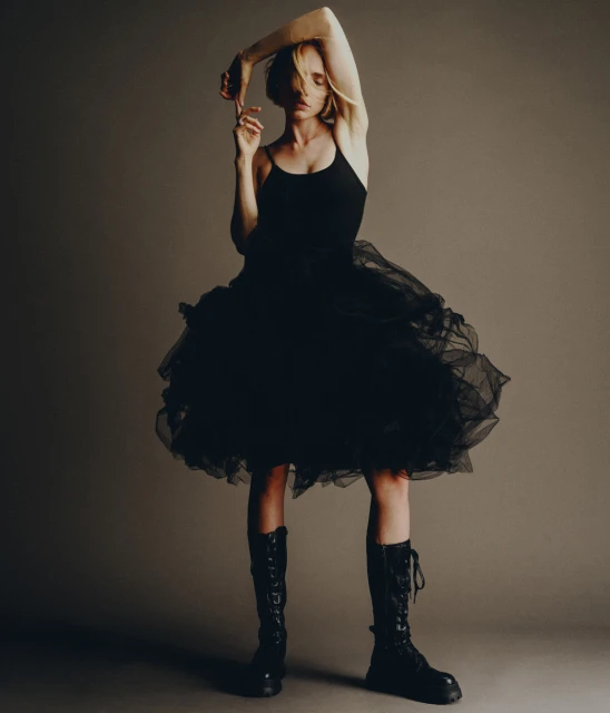 Fashion shot of a model in a black dress and combat boots, standing in a photo studio with arms raised, against a neutral background.