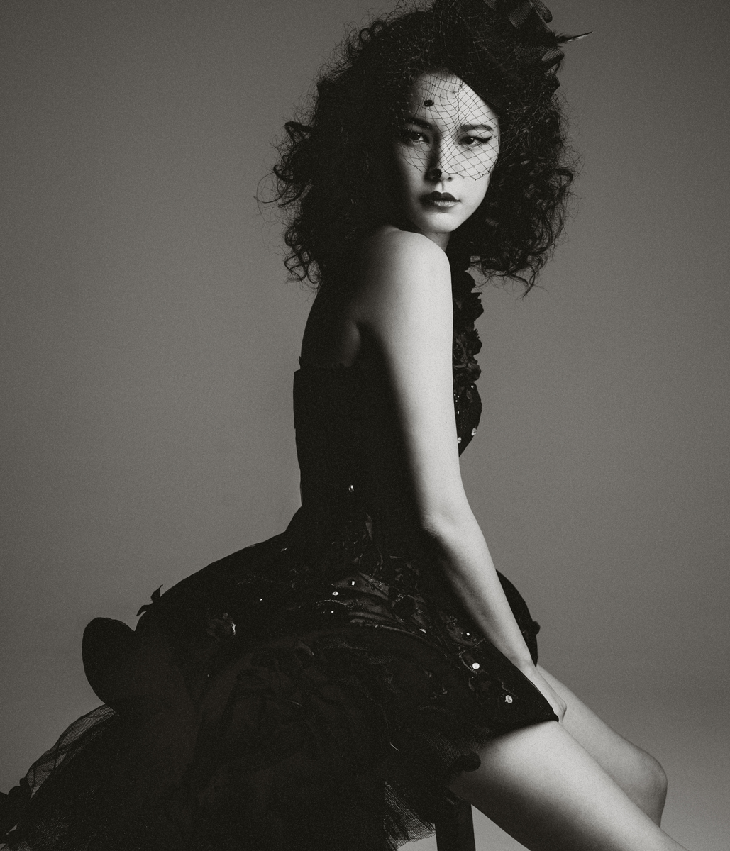 Portrait of an adult female model wearing a black dress with a veil, posing in a studio with neutral background and soft lighting.