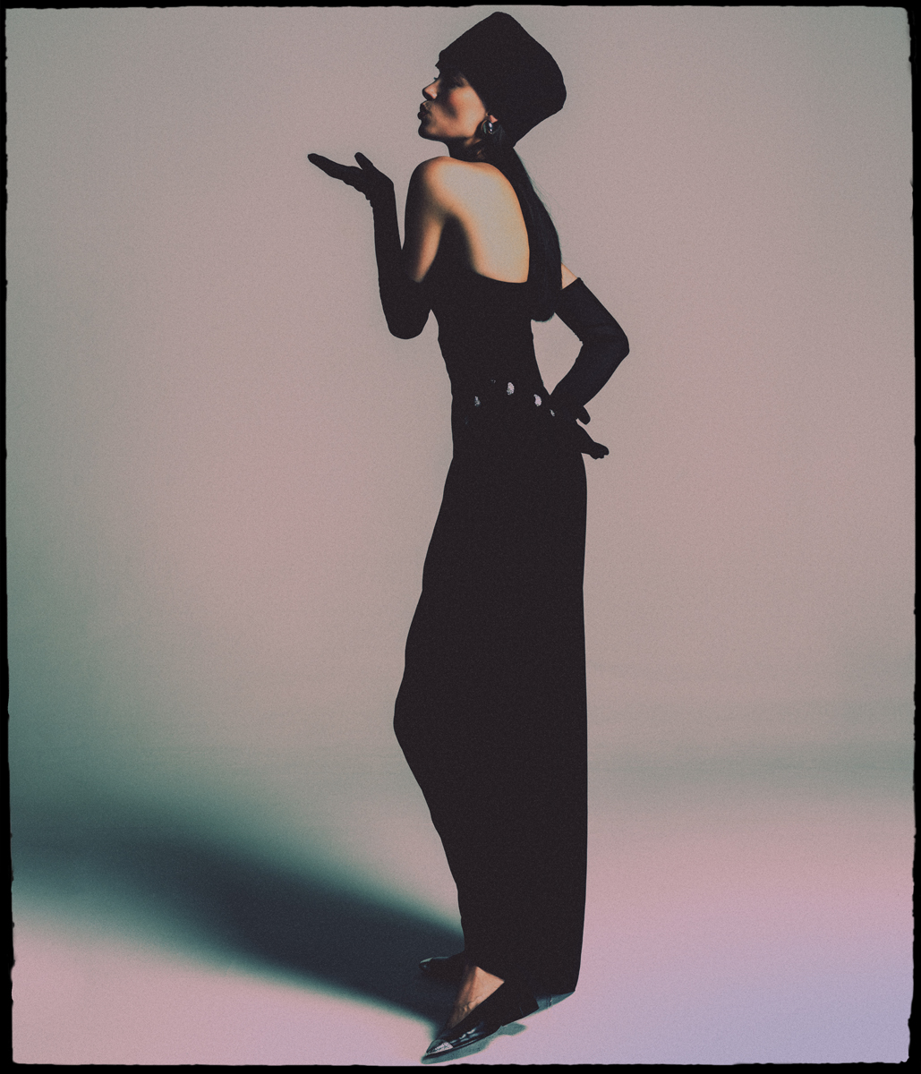 Elegant woman in black hat and gown blowing a kiss in a fashion shot, studio lighting, fashionable, stylish and graceful.