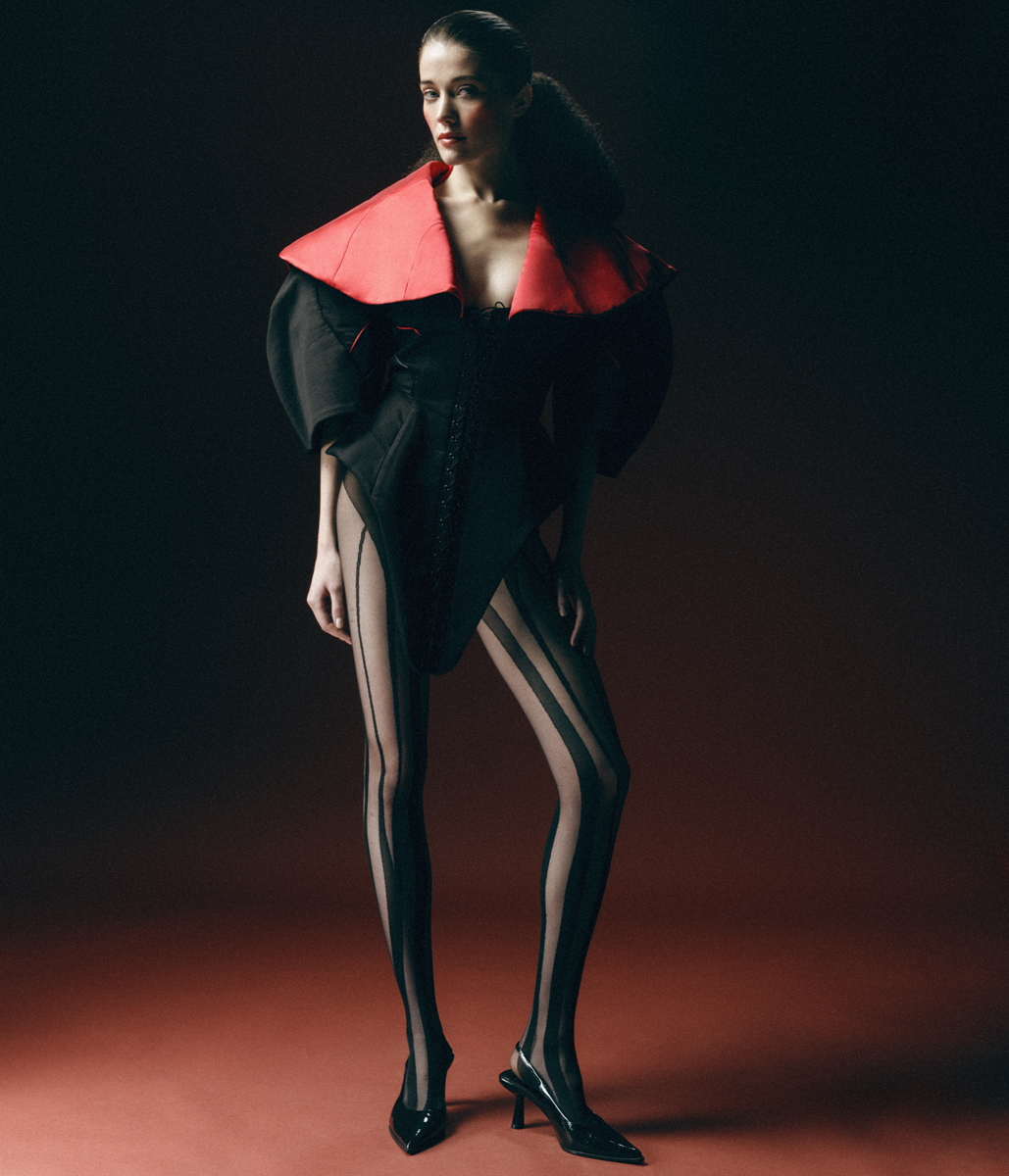 Fashion model in a black bodysuit with a red collar and black striped tights poses against a dark red gradient background.