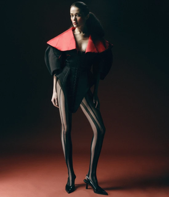 Fashion model in a black bodysuit with a red collar and black striped tights poses against a dark red gradient background.