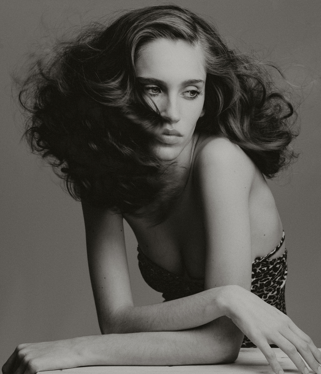 Monochrome portrait of a young woman with tousled brown hair, wearing a patterned top, striking a pose against a grey background.