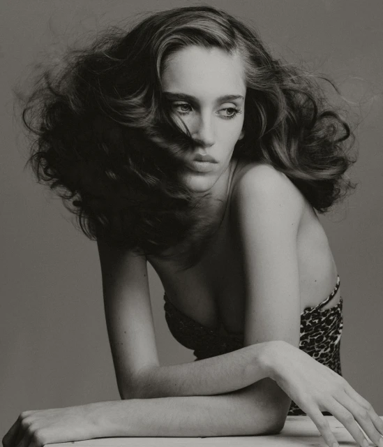 Monochrome portrait of a young woman with tousled brown hair, wearing a patterned top, striking a pose against a grey background.
