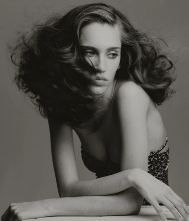 Monochrome portrait of a young woman with tousled brown hair, wearing a patterned top, striking a pose against a grey background.