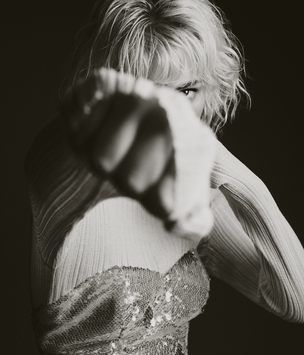 Portrait of a confident adult woman with blonde hair, wearing a shimmering top, her fist close to the camera, indoor in dark setting.