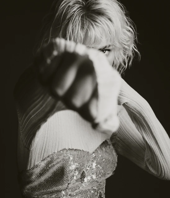 Portrait of a confident adult woman with blonde hair, wearing a shimmering top, her fist close to the camera, indoor in dark setting.