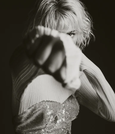 Portrait of a confident adult woman with blonde hair, wearing a shimmering top, her fist close to the camera, indoor in dark setting.