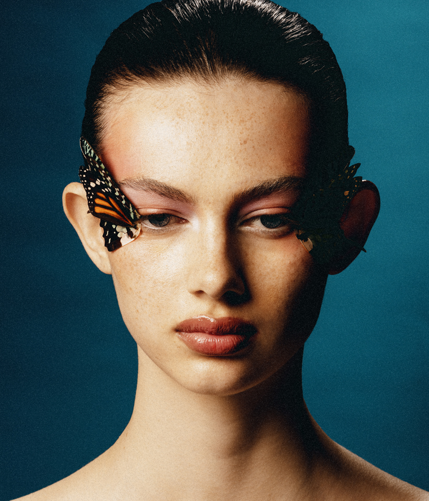 Portrait of a woman with butterfly wing accessories, featuring pink eyeshadow and a dark blue background, for beauty or fashion visuals.