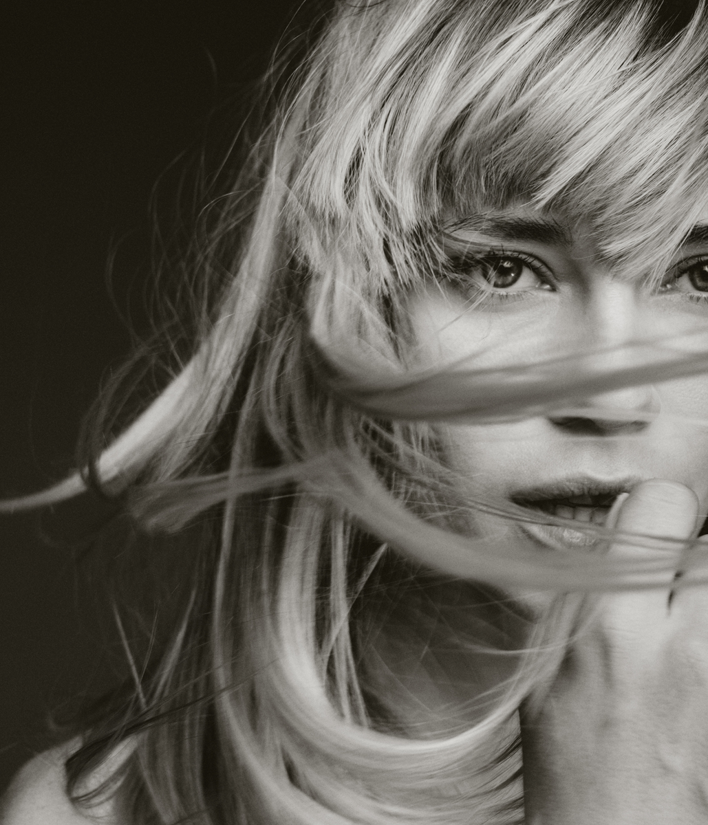 Monochrome portrait of a blonde woman with windblown hair covering part of her face, emphasizing her eyes and serene expression.