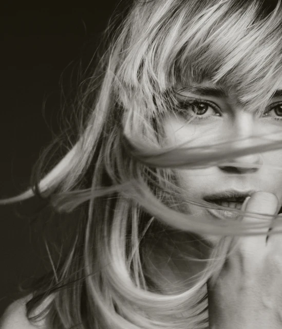 Monochrome portrait of a blonde woman with windblown hair covering part of her face, emphasizing her eyes and serene expression.