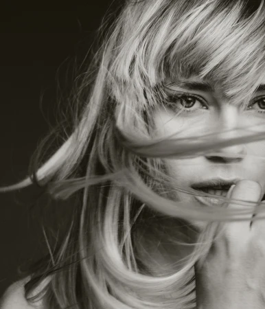 Monochrome portrait of a blonde woman with windblown hair covering part of her face, emphasizing her eyes and serene expression.