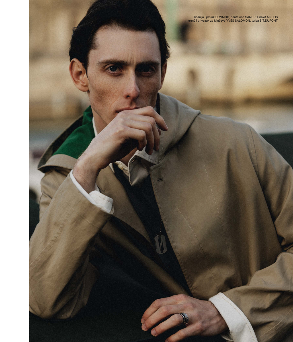 Adult male with hand on face, wearing a trench coat and ring, posing outdoors with blurred background.