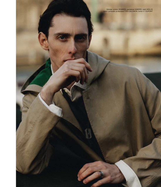 Adult male with hand on face, wearing a trench coat and ring, posing outdoors with blurred background.