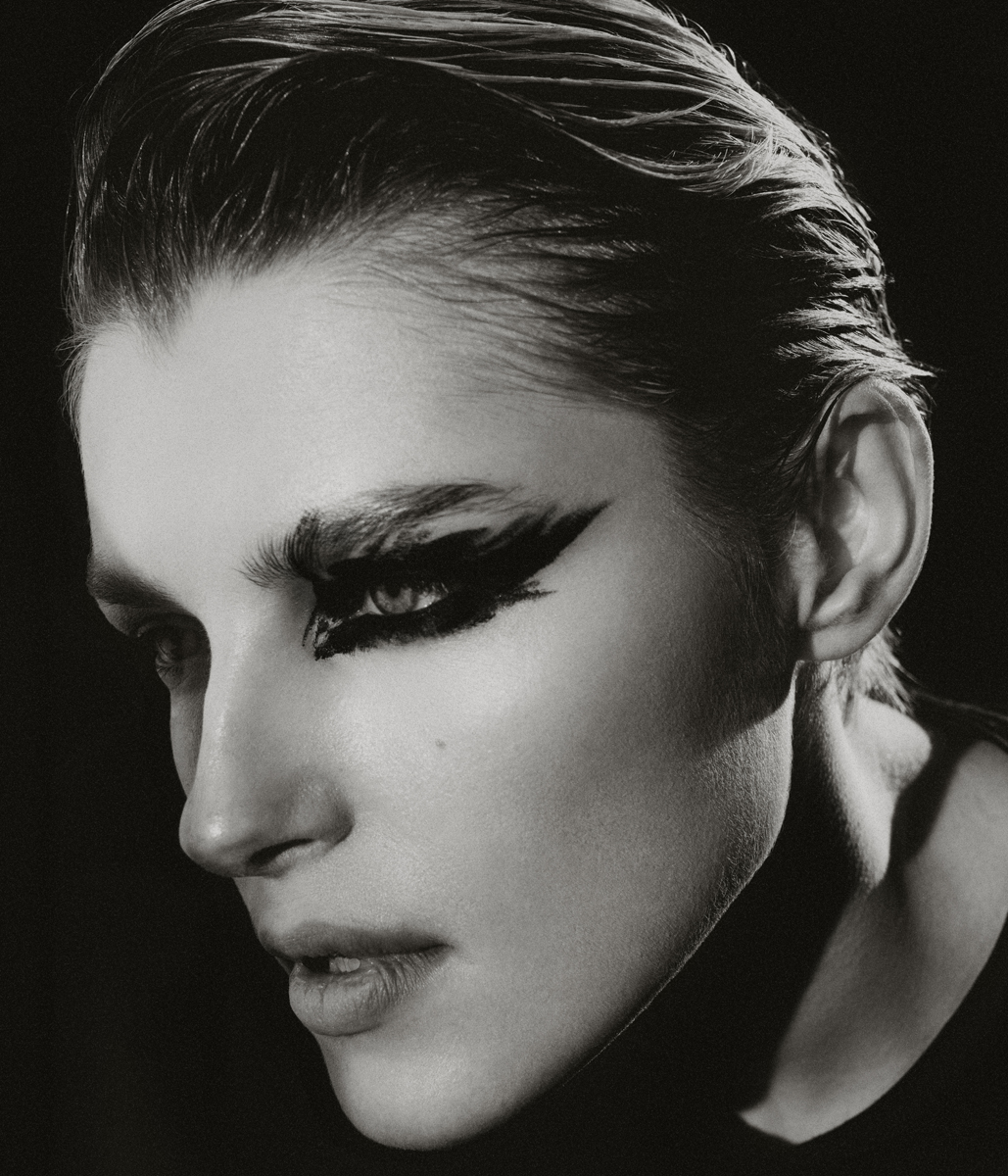 Monochrome portrait of a young adult woman with dramatic black eye makeup in a close-up studio shot against a dark background.