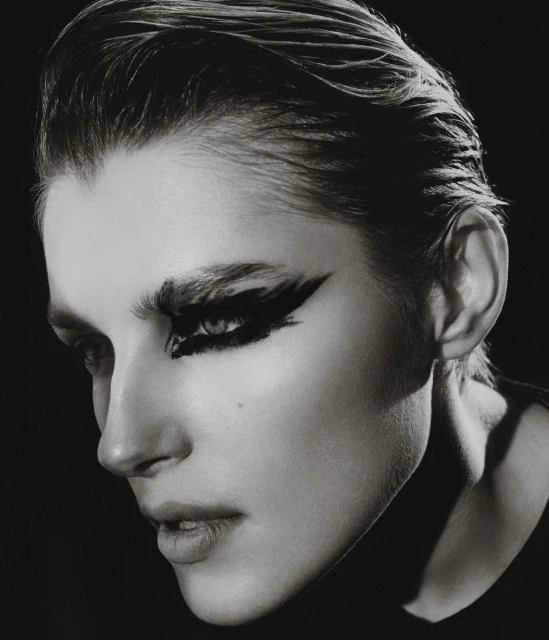 Monochrome portrait of a young adult woman with dramatic black eye makeup in a close-up studio shot against a dark background.