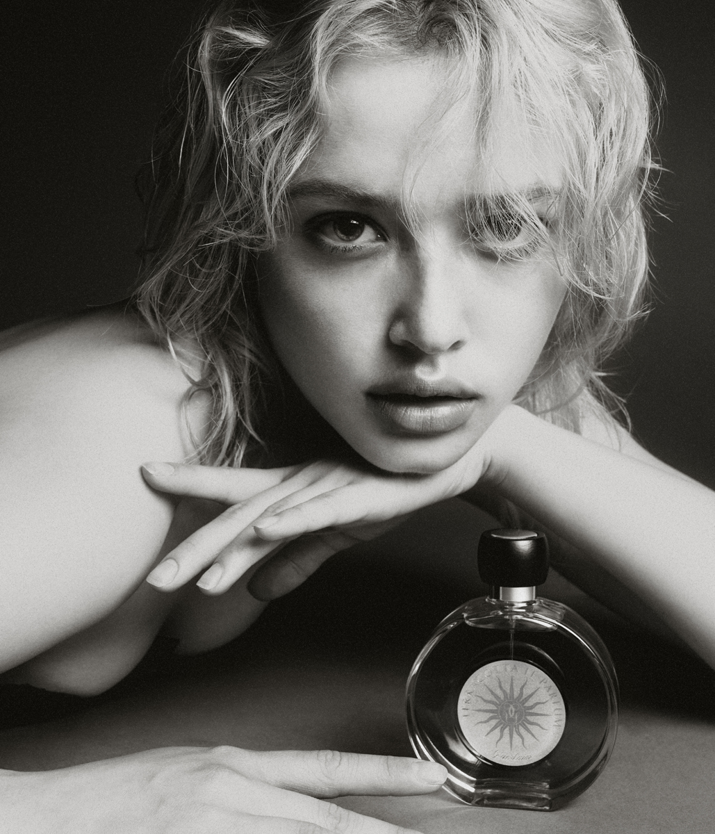 Black and white portrait of a blonde woman with a perfume bottle, resting her face on her hands in a close-up indoor shot.