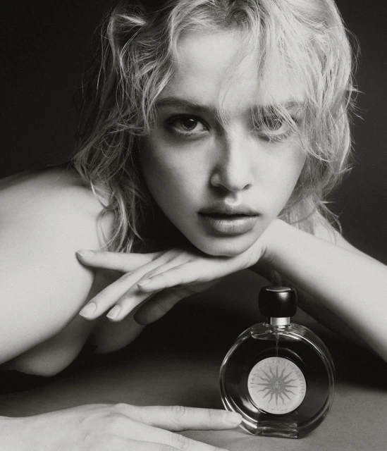 Black and white portrait of a blonde woman with a perfume bottle, resting her face on her hands in a close-up indoor shot.