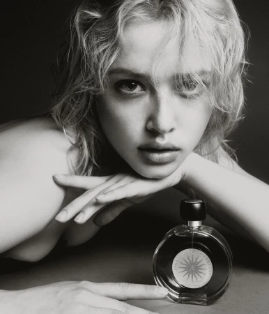 Black and white portrait of a blonde woman with a perfume bottle, resting her face on her hands in a close-up indoor shot.