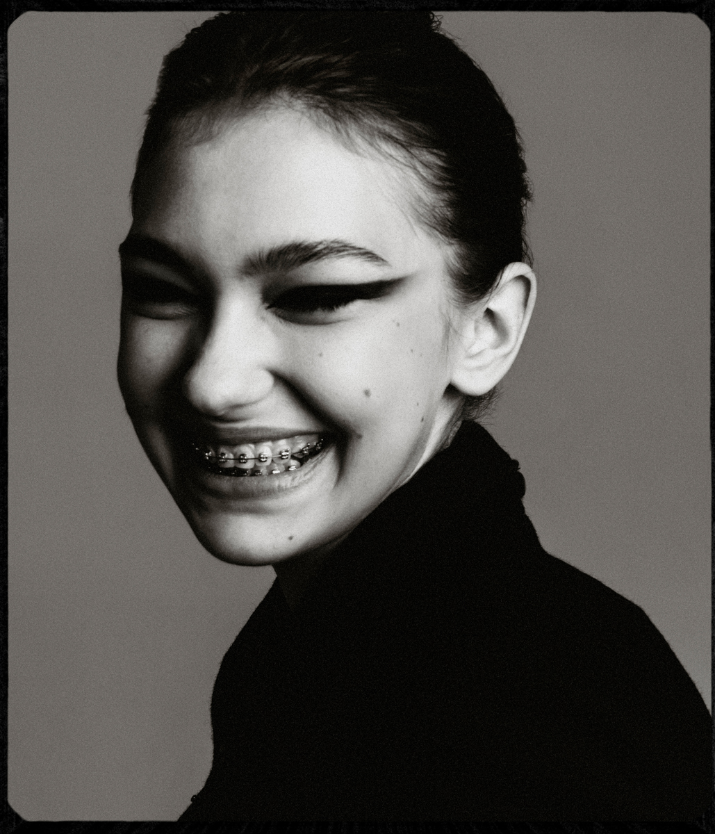 Black and white portrait of a smiling young woman with braces, wearing a black turtleneck sweater against a plain grey backdrop.