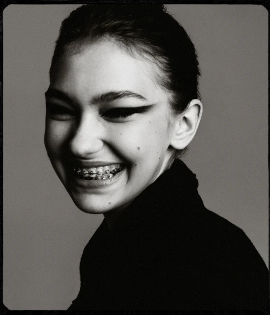Black and white portrait of a smiling young woman with braces, wearing a black turtleneck sweater against a plain grey backdrop.