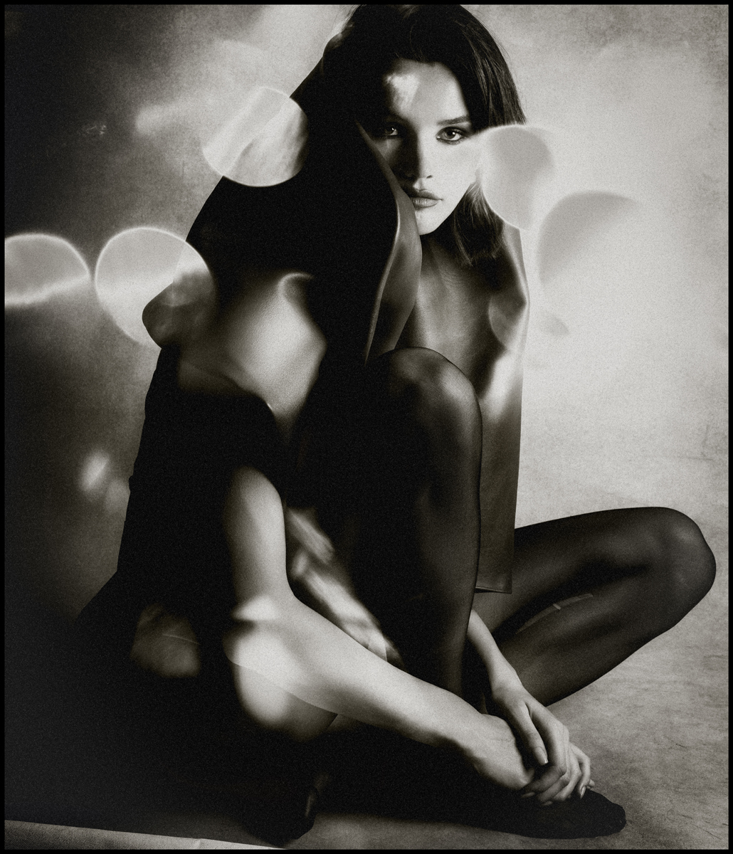 Monochrome portrait of a woman sitting cross-legged. She has dark hair and a thoughtful expression, in an artistic, studio setting.