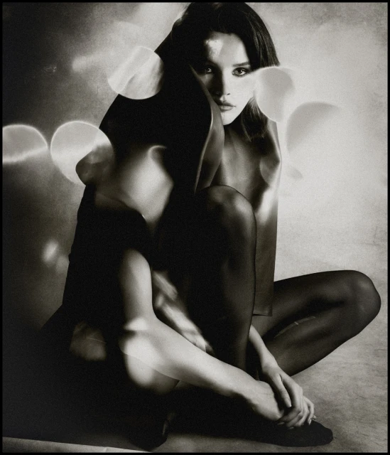 Monochrome portrait of a woman sitting cross-legged. She has dark hair and a thoughtful expression, in an artistic, studio setting.