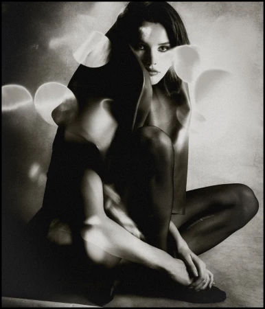 Monochrome portrait of a woman sitting cross-legged. She has dark hair and a thoughtful expression, in an artistic, studio setting.