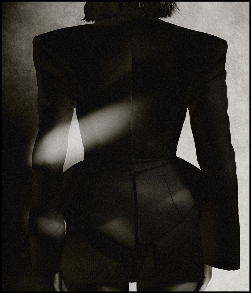 Back view of person wearing black jacket with dramatic shoulders in a studio setting. The image is in black and white.