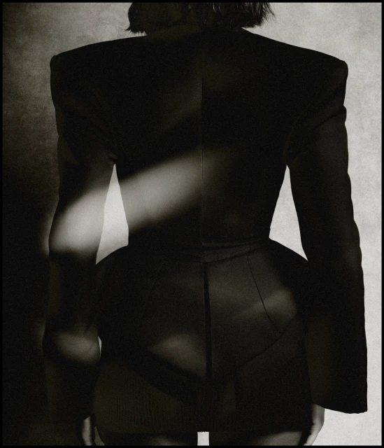 Back view of person wearing black jacket with dramatic shoulders in a studio setting. The image is in black and white.