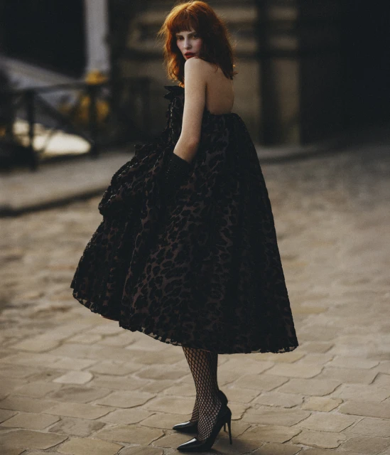 Adult woman with red hair poses in a black floral dress, black fishnet stockings, and black heels on a tan cobblestone street.