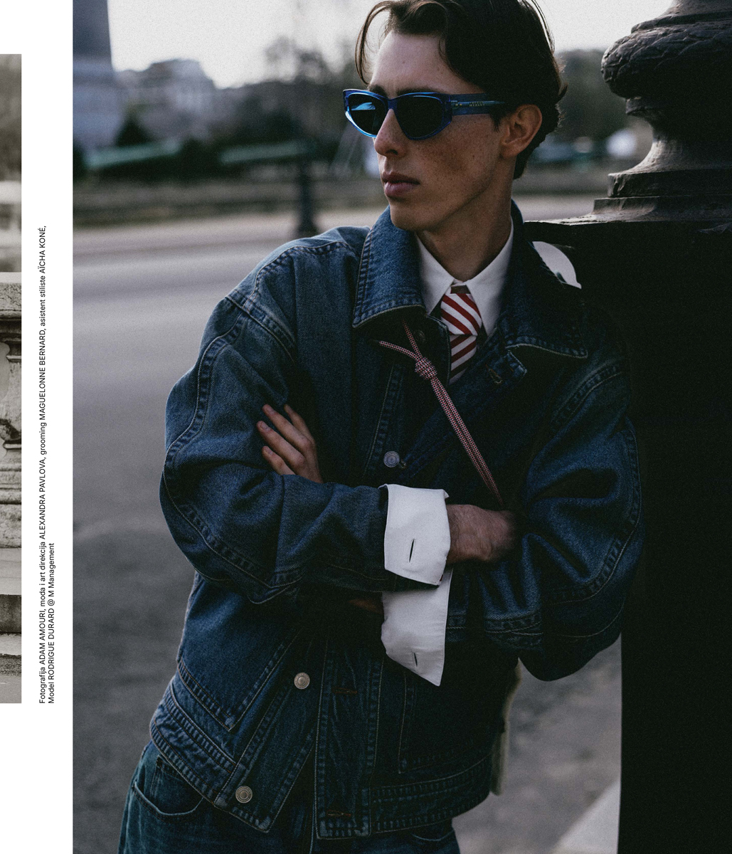 Fashionable young adult male posing outdoors. He is wearing a denim jacket, a white collared shirt, tie, and blue sunglasses.