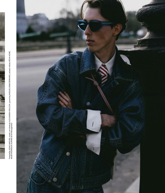 Fashionable young adult male posing outdoors. He is wearing a denim jacket, a white collared shirt, tie, and blue sunglasses.