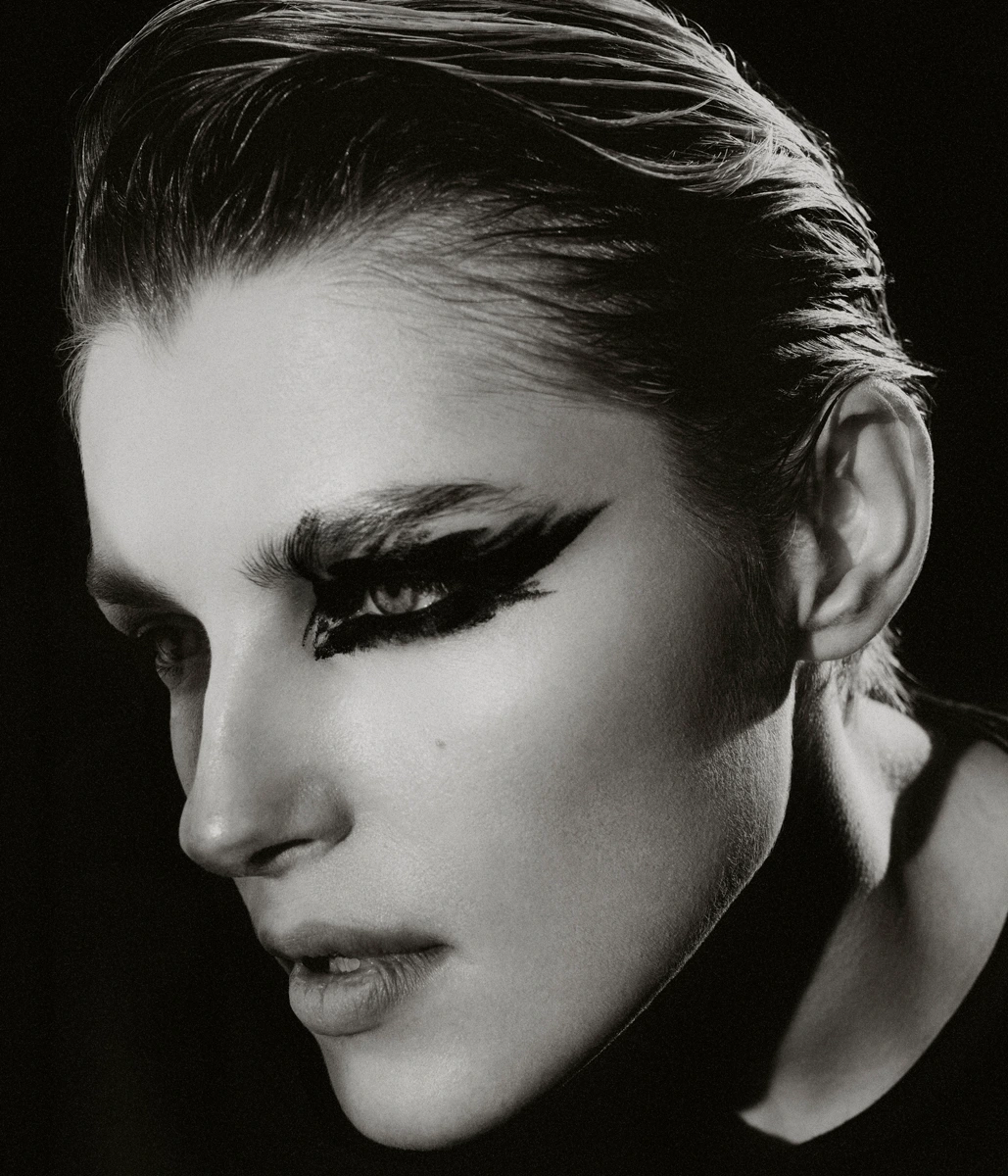 Black and white close-up portrait of a model with slicked-back hair and dramatic feathered black eye makeup against a dark background.