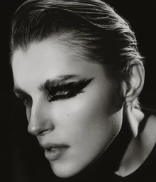 Black and white close-up portrait of a model with slicked-back hair and dramatic feathered black eye makeup against a dark background.