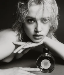 Black and white portrait of a woman resting her chin on her hand beside a round glass perfume bottle against a dark background.