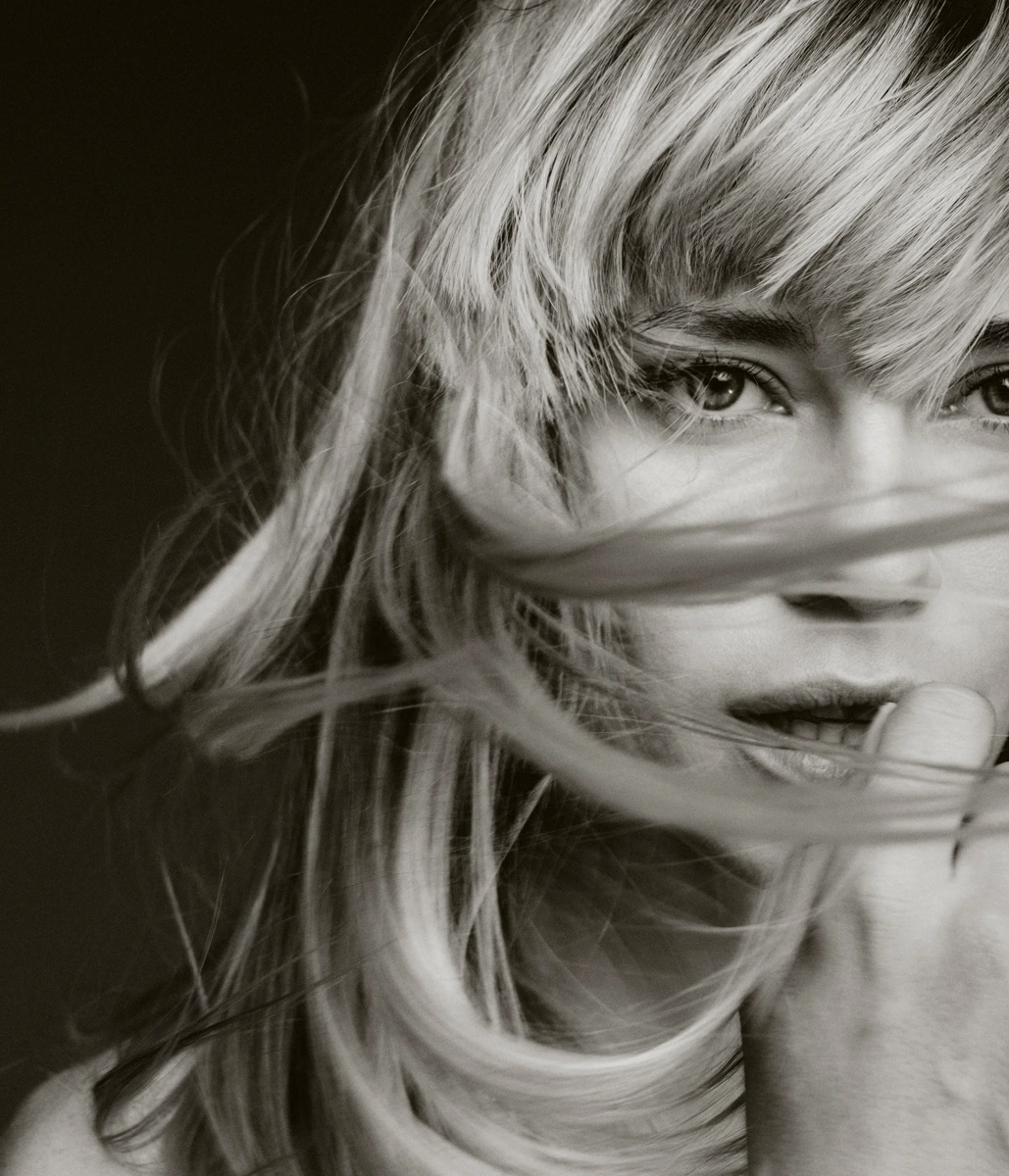Monochrome close-up portrait of a woman with fringe bangs and strands of hair blowing across her face against a dark background.